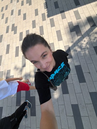 A high-angle, full-body selfie is captured on a paved walkway in Belaya Kholunitsa, Russia. The walkway is constructed with alternating light and dark rectangular stone pavers arranged in a staggered pattern. Sunlight casts shadows of nearby trees onto the ground. The central figure, a woman with dark hair pulled back, is wearing a black t-shirt with a vibrant blue graphic design and a brown strap across her shoulder. She is smiling and looking towards the camera, holding the camera with her right arm, which is extended. In the foreground, to the left of the central figure, two other individuals are visible. One person is wearing a white t-shirt and red shorts, with their legs bent as if walking or running. The other individual, closer to the bottom of the frame, is wearing black pants and dark shoes, with their legs also appearing to be in motion. The overall impression is of people walking outdoors on a sunny day.
