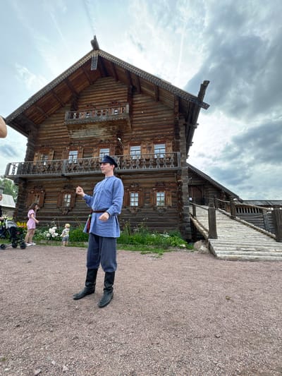 In Nevskii Parkleskhoz, Russia, a person dressed in traditional clothing stands in front of a large, ornate wooden building. The building is constructed from logs and features intricate carvings and balconies. A wooden ramp leads up to an entrance on the right side of the building. The ground is covered with gravel, and there are some plants and flowers visible. The sky is cloudy. Other people are present in the background, including a woman with a stroller and a child.