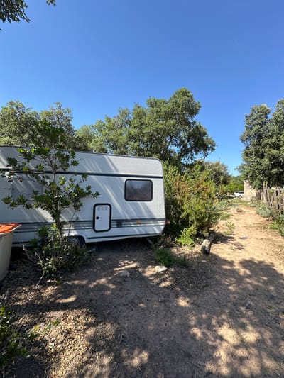 A static shot captures a white caravan parked on a dirt path bordered by dense green trees and brush under a clear blue sky. The caravan is positioned on the left side of the frame, with its side facing the viewer, revealing a window and a closed door with a vent. A small tree with green leaves grows in front of the caravan. To the right of the caravan, the path curves away, leading past a rustic wooden fence. In the distance, a white vehicle is visible. The ground is covered with dirt, dry leaves, and scattered stones, with dappled sunlight creating shadows from the foliage. The scene appears to be in an outdoor, possibly rural, setting, consistent with the description of Santa Cristina d'Aro, Spain, a town known for its natural surroundings and proximity to the Mediterranean coast. There are no people or animals visible in the image.