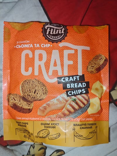 The image is a close-up of a package of Flint Craft Bread Chips with salmon and cheese flavor, presented on a surface that appears to be a bed or a blanket with a red and white patterned textile. The packaging is predominantly orange with white lettering and features an image of grilled salmon, cheese slices, and rye bread slices. The Flint logo is visible at the top, along with the text "CRAFT BREAD CHIPS" in black rectangles. The bottom section of the packaging displays nutritional information, a depiction of the chips opening, and an illustration of a hammock. The location of Skorokhodove, Ukraine, is not evident in this image.