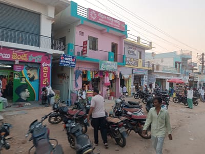 An outdoor scene in Kothacheruvu, India, depicts a busy commercial street with multi-story buildings and numerous individuals. The time of day appears to be late afternoon or early evening under a clear, light-colored sky. The street is an unpaved dirt road with scattered debris.
On the left, a white building with a gray roll-down shutter is visible above a pink storefront with a sign in Telugu advertising "Sai Gents Salon," featuring images of men's hairstyles. Inside and outside this establishment, approximately five men are present, some seated.
In the center, a pink building with teal trim and a balcony bears a prominent sign for "KEERTANA GOLD LOANS" in both English and Telugu. Below this, a shop displays various items of clothing on hangers. To the right of this building, a light gray/yellow building with a "MADHU Tailors" sign is visible.
Numerous motorcycles and scooters are parked haphazardly along both sides of the street, with several visible in the foreground and midground. One man, wearing a light green long-sleeved shirt and light pants, walks towards the viewer in the foreground, right of center. Another man, dressed in a light beige t-shirt and dark pants, walks away from the viewer in the midground, between parked motorcycles.
Further down the street on the right, a red market umbrella is present, and several individuals are standing near shops. A man in a white shirt and green lungi stands near the umbrella. Overhead power lines crisscross the sky, and utility poles with tangled wires are visible on the right side of the street. Approximately 15-20 individuals are present throughout the scene, engaged in various actions such as walking, standing, and sitting, indicating a public gathering or commercial activity.