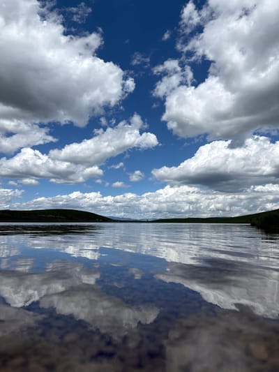 The image presents a serene natural landscape located in Javorje, Montenegro, captured from a very low-angle perspective near the water's edge. The upper portion of the frame features a deep blue sky populated by numerous large, white cumulus clouds, many with grey undersides, indicating significant vertical development. Below the horizon line, which is subtly undulating, a expanse of calm water reflects the sky and clouds with notable clarity, the reflections slightly distorted by gentle ripples on the surface. Distant, low-lying green hills or mountains define the landscape along the horizon, appearing sparsely vegetated. The immediate foreground, at the bottom of the image, shows the shallow water and indistinct rocky or sediment details on the bottom. There are no people, animals, structures, or artificial objects visible within the scene. The environment appears pristine and undisturbed under bright natural daylight.