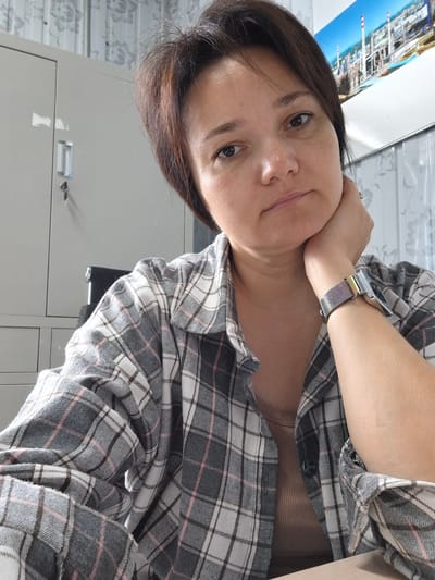An indoor, medium shot shows a woman with short, dark brown hair, resting her chin on her right hand. She is wearing a grey and white plaid shirt over a light brown tank top. A watch with a patterned band is visible on her left wrist. Behind her, a light grey metal cabinet is on the left, and a patterned curtain is on the right. A framed picture of an industrial complex is mounted on the wall above the curtain. The image appears to be taken within the Tecnimont camp in Russia.