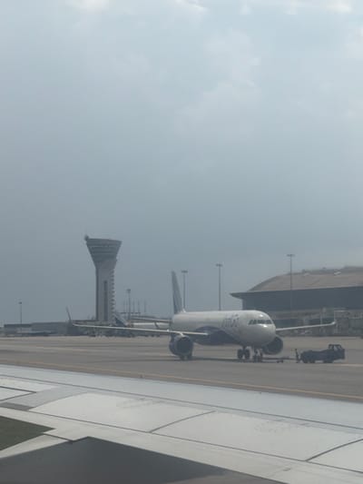 This image, taken at Rajiv Gandhi International Airport in Mamidipally, India, captures an aerial view of an airport tarmac on a hazy day. In the foreground, the wing of a commercial airplane is visible, extending from the left side of the frame. Further back on the tarmac, a white Indigo Airlines passenger jet is parked, its nose facing towards the right. The jet has its landing gear deployed, and a ground service vehicle, possibly a baggage tug, is positioned near its rear. To the left of the jet, a distinctive control tower stands tall, characterized by its unique, tapering design and numerous windows. In the background, a long, low-rise airport building with a curved roof is partially visible. The sky is overcast with diffuse light, and the overall atmosphere is muted due to the hazy conditions.