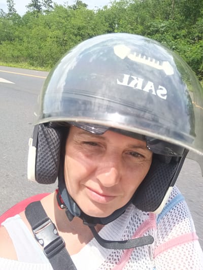 A close-up photo shows a woman wearing a motorcycle helmet, driving along a paved road with green trees and vegetation visible in the background. The helmet is black with a clear visor, and the word "SAKL" is printed on the front, backwards. She is wearing a white top with pink and blue stripes, and a black strap is fastened across her chest. The road appears to be in Plai Phraya, Thailand.