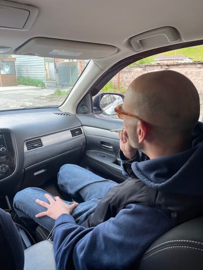 A man with a shaved head is sitting in the passenger seat of a car, looking out the window and smoking. He is wearing a navy blue hoodie and blue jeans. The car's interior is visible, including the dashboard, steering wheel, and center console. The exterior of the car shows a street scene with buildings and greenery. The location is Arsha, Georgia.