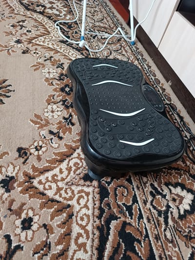 A black vibrating platform for fitness is positioned on a patterned carpet indoors. The platform has a textured surface with small raised circles and two curved white lines. A white electrical cord is visible connected to a stand in the background. The carpet features a traditional floral and swirling design in shades of brown, beige, and black. The overall scene suggests a home or gym environment. The location is Vuhlehirsk, Ukraine.