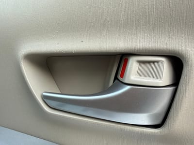 The image shows a close-up view of a car's interior door handle. The handle is made of a metallic silver material and has a sleek, ergonomic design. It is set against a textured beige plastic panel, which is characteristic of a car's interior door trim. A small, rectangular button with a red insert is visible on the left side of the handle, likely for unlocking or child safety. The lighting suggests an indoor setting, possibly a garage or showroom, as there are no strong shadows or indications of natural light. The overall impression is one of a well-maintained, functional car interior. There is no text visible in the image.