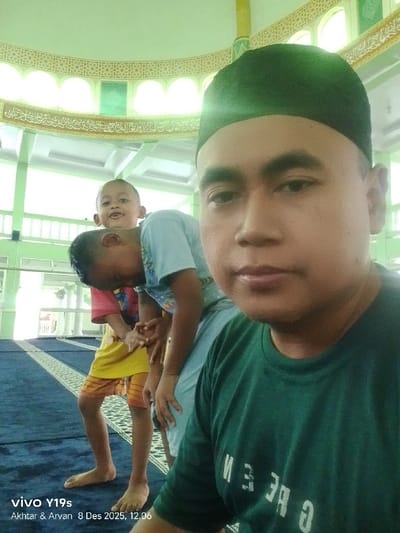 Inside a mosque, a man wearing a green cap and T-shirt takes a selfie. Two children are standing behind him, one wearing a blue shirt and the other in pink and yellow attire. They all stand on a blue patterned carpet. Ornate architectural details adorn the mosque's interior, including arched windows, decorated ceilings, and pillars. The image's timestamp indicates it was taken on December 8, 2025.