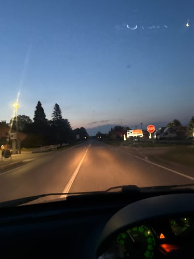 The image depicts a driver's perspective from inside a vehicle traveling on a road at dusk. The sky is a deep blue, transitioning to lighter hues near the horizon, with a few faint clouds visible. The road ahead is illuminated by the vehicle's headlights and streetlights along the side, creating a warm glow. Trees and buildings line the road, casting shadows in the low light. A red octagonal stop sign is visible on the right side of the road, along with illuminated billboards and what appear to be parked vehicles. The interior of the vehicle is partially visible, with the dashboard and parts of the steering wheel in the foreground, including illuminated gauges. The scene appears to be set in a semi-rural or suburban area outside the city of Ponteterra, Italy.