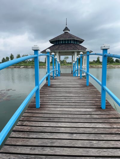 The image shows a wooden pier with bright blue railings leading towards a gazebo on a calm lake. The sky is overcast with gray clouds. Trees and vegetation line the distant shore. The gazebo has a multi-tiered roof and white pillars. The pier is constructed of weathered wooden planks. The lake has lily pads visible on its surface. The location is identified as Severynivka, Ukraine.