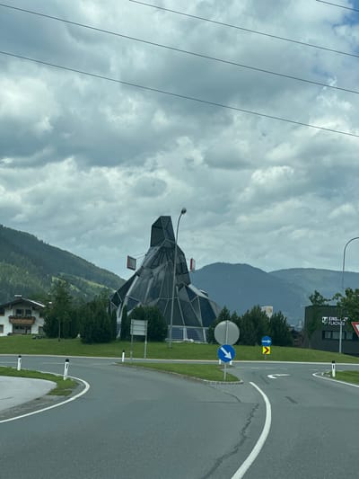 This media file captures a view from a vehicle, likely moving along a road, in a mountainous region. The foreground is dominated by a wet asphalt road with white lane markings, indicating a recent or ongoing rainfall. To the left of the road, a grassy verge is visible, lined with trees and a striking, modern building with a faceted, geometric glass exterior. Solar panels are mounted on the building. Further back, dense, dark green pine-covered mountains rise steeply, their peaks obscured by heavy, overcast clouds. The sky is a mix of grey and white, suggesting a cloudy or stormy day.
On the grassy verge, several traffic signs are present. One circular blue sign with a white arrow pointing right is mounted on a pole, and another, a blue square sign with a yellow triangle containing an upward-pointing arrow, is also visible. A lamppost arches over the scene from the left. In the background, a building with the partially visible text "EINS... ZF... FLACHAU" can be seen, providing a location clue. The overall impression is one of a natural landscape dramatically contrasted with modern architecture and infrastructure, under a somber sky. The scene appears to be set in the daytime, though the weather conditions cast a dim light. There are no people visible in the frame.