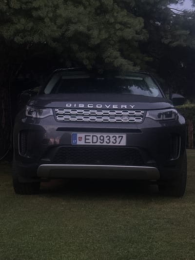 A dark gray Land Rover Discovery Sport is parked on a green lawn, facing the camera. The vehicle is positioned beneath a large, dark green conifer tree, with its branches casting shadows over the car. The front of the car is visible, including the headlights, grille with the "DISCOVERY" emblem and Land Rover logo, and a license plate that reads "ED9337" and has the "LT" country code. The car appears to be in good condition. The background shows more of the conifer tree and hints of a fence and some foliage. The lighting suggests it is either early morning or late evening. No people are visible in the image. The location is identified as Lapės, Lithuania.