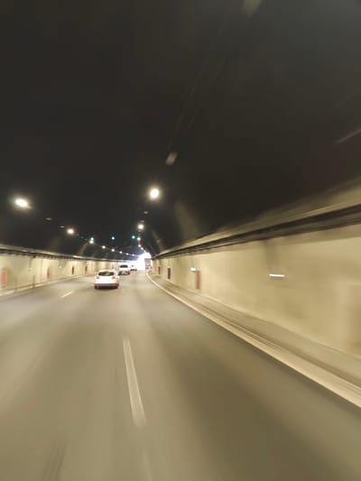 The image shows a view from inside a tunnel, likely a highway tunnel given the presence of multiple lanes and vehicles. The tunnel has a dark, arched ceiling with numerous lights illuminating the interior. The walls of the tunnel are a light, neutral color, with some recessed lighting and what appear to be red emergency call boxes visible.
Several vehicles are present on the road, moving away from the viewer and towards the light at the end of the tunnel. The closest vehicle is a white SUV or hatchback, followed by other vehicles further ahead, including what appears to be a white passenger car and a truck. The motion blur indicates that the image was taken while in motion, likely from a moving vehicle. The road surface is asphalt with visible lane markings.
The context provided about the location, Gorno Kamartsi, Bulgaria, suggests this is a public infrastructure scene. The overall impression is one of travel through an enclosed, man-made passage.