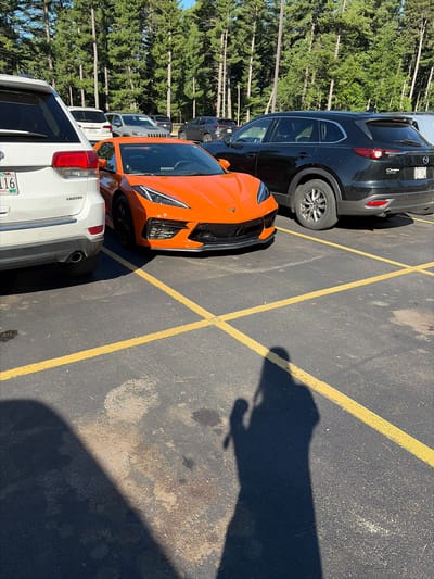 A high-angle, medium shot captures a row of parked cars on an asphalt lot in Wisconsin Dells, Wisconsin, USA. The foreground is dominated by the shadow of a person holding a camera, cast across the yellow parking lines and dark pavement. To the left, the rear of a white Jeep Grand Cherokee is visible, its red taillight and "LIMITED" badging prominent. Adjacent to the Jeep, a vibrant orange Chevrolet Corvette C8 is parked, its sleek design and sharp angles clearly defined. Behind the Corvette, a black Mazda CX-9 SUV is parked, its rear bumper and taillight visible. In the background, a dense forest of tall, green pine trees rises against a clear blue sky. The scene appears to be a sunny day in a parking lot, likely near a tourist area given the variety of vehicles.