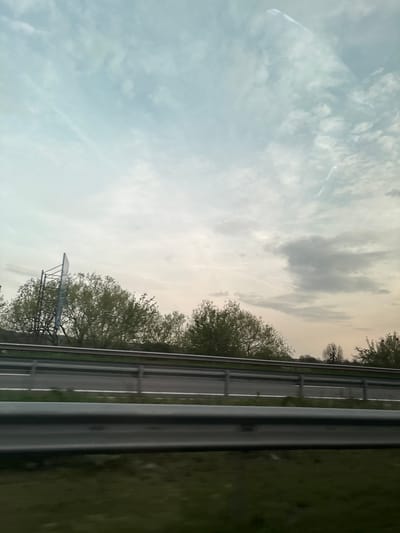 The image depicts a view from inside a moving vehicle on a highway, likely in or near Maasmechelen, Belgium, though specific landmarks are not visible. The scene includes a multi-lane highway with a metal guardrail dividing the roadway from a grassy area. Trees line the side of the road. The sky is overcast with visible clouds. The photograph is taken through a window, and a portion of the vehicle's interior is visible. A road sign with its frame is barely visible from the side.