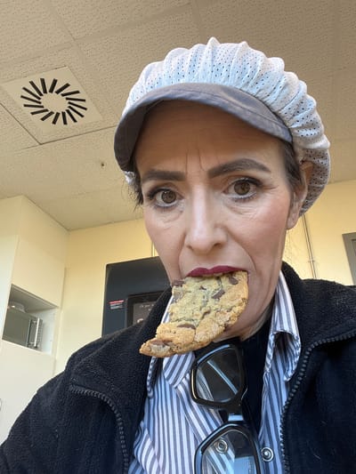 A woman is depicted in a selfie, holding a chocolate chip cookie in her mouth. She is wearing a white hairnet, a blue and white striped shirt, and a black fleece jacket. A pair of black-rimmed glasses are visible next to the cookie. The background includes a ceiling with a circular air vent, light yellow walls, and a black electronic device mounted on the wall above white cabinets. The location is identified as Mensdorf, Luxembourg.