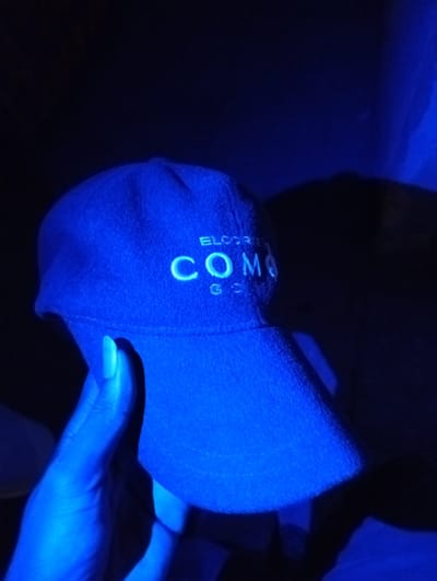 A hand holds a dark blue baseball cap under intense blue light. The cap has white embroidered text that appears to read "ELCORTE" above a larger "COMO" and possibly "GO" below. The background is dark, with some faint details suggesting fabric or shadows. The hand holding the hat is also illuminated with the same blue light.