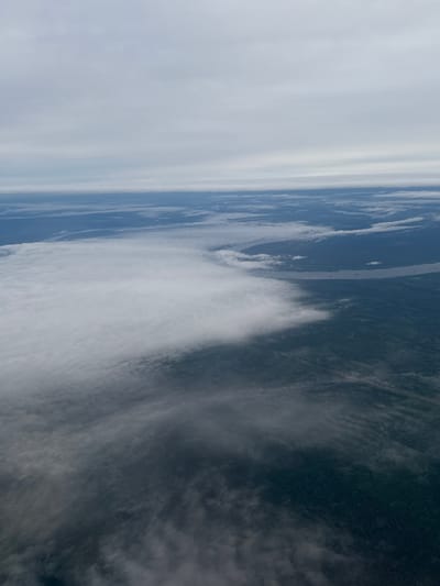 This aerial image shows a river winding through a landscape in the vicinity of Yugo-Kamskiy, Russia. A large patch of white, possibly clouds or fog, dominates the lower right portion of the frame. The river snakes through a wooded area before disappearing into the distance. The sky is overcast.