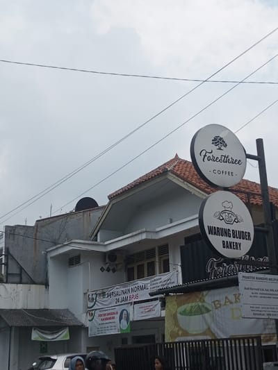 The image captures a street scene dominated by buildings and signage under an overcast sky. The sky, a uniform light grey, occupies the upper third of the frame, crisscrossed by several black power lines.
In the mid-ground, a white two-story building with a distinctive reddish-brown tiled roof is prominent on the right. Its architecture includes intricate eaves and what appears to be an air conditioning unit mounted on the wall. Below its windows, a large white banner is displayed, featuring text in Indonesian that reads "PERSALINAN NORMAL, 24 JAM BPJS" (Normal Delivery, 24-hour BPJS), indicating a medical or maternity service. Beneath this, another banner reads "DOKTER SPESIALIS KANDUNGAN" (Obstetrician/Gynecologist Specialist) and shows a picture of a male doctor. To the left of this building, a taller, grey structure with a flatter roof and a dark satellite dish on top is visible. Further to the far left, a lower structure with a corrugated metal roof is partially seen.
On the right side of the image, two circular white signs are stacked vertically on a black pole. The top sign has a tree logo and reads "Foresthree - COFFEE -", suggesting a coffee shop. The bottom sign displays a chef hat logo with a cartoon face and reads "WARUNG BLUDER BAKERY", indicating a bakery. Below these signs, a dark-colored storefront or awning is partially visible, with "Foresthree" text, likely marking the entrance to the combined coffee shop and bakery. To the right of these signs, a smaller banner indicates a "PRAKTEK MANDIRI AKUPU" (Independent Acupuncture Practice) run by "Budi Darmadi S.Si, Apt, MM, Am", with a phone number and consultation hours.
At the very bottom of the image, parts of people's heads are visible, suggesting they are on a street or sidewalk, partially obscured by a black metal fence or gate. A white car is also partially visible behind the fence on the left. The overall lighting suggests it is daytime, under cloudy or overcast weather conditions. The language on the signs and banners indicates the scene is likely in Indonesia.