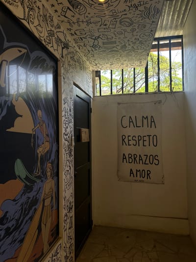 The image shows a narrow hallway with white walls and a tiled floor. The hallway is located in Cobano District, Costa Rica, at latitude 9.629 and longitude -85.152. A black door is on the left side of the hallway. The door is closed. To the right is a white canvas sign with black lettering that reads "CALMA RESPETO ABRAZOS AMOR" and is signed "AJ. ARTE". Above the sign is a barred window that looks out onto a grove of trees. The hallway is decorated with black and white drawings that cover most of the walls. One drawing depicts a skeleton riding a surfboard on a wave. Another drawing shows a woman wearing a swimsuit. The image is taken at a slightly downward angle. The hallway is dimly lit. There are no people in the image.