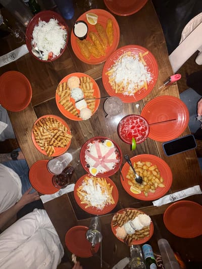 A high-angle shot depicts a wooden table laden with orange-red plates of food and beverages. The setting appears to be a restaurant, with portions of people visible around the edges of the frame. There are no distinguishing characteristics to allow the determination of the location in Des Plaines, United States of America.
Various dishes are arrayed across the table: one plate contains grilled pita bread; another contains fried potatoes topped with grated white cheese; a third has fried potatoes with a lemon wedge; another plate has salad with shredded white cheese; a plate has fried fish strips with a dipping sauce and a lemon wedge; another has a dish containing a pink cocktail. There are multiple empty plates also visible. Additional objects include a glass of water, a glass of dark liquid, a smartphone, a bottle of sparkling water, silverware, and a pink cigarette lighter. Visible partial people include those wearing light-colored shirts and jeans, with a tattoo visible on an arm.