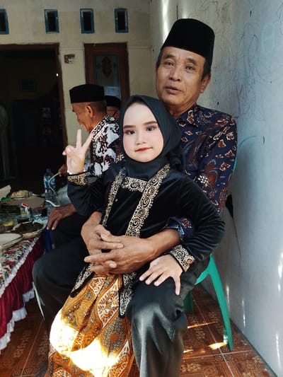 In Juwana, Indonesia, an image features an adult male and a child seated in a semi-outdoor setting. The adult male, on the right, wears a black peci and a dark blue batik shirt, and is seated on a light green plastic chair. He holds a child on his lap, with his hands clasped over the child's left leg. The child, centrally positioned, wears a black hijab and a black velvet outfit adorned with gold and white embroidery, and has visible facial makeup. The child's right hand forms a "V" sign, while the left hand rests on the adult's leg. Both subjects in the foreground look directly forward. To the left in the midground, an elderly male, wearing a black peci and a patterned batik shirt, is seated facing away from the camera, partially obscured. A low table, covered with a white and red frilly tablecloth, holds plates, a plastic water bottle, and wrapped food items. The background includes a light-colored wall with faint markings on the right, and a light brown wooden door with three rectangular openings above it on the left. The floor is composed of patterned ceramic tiles. Direct sunlight illuminates portions of the scene, casting distinct shadows.
