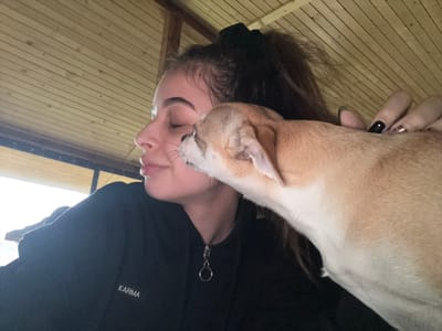 A close-up image depicts a young female and a small dog within an indoor setting in Ralevo, Bulgaria. The female, positioned on the left, is shown in profile, facing left, with her eyes closed or partially closed. She has dark hair styled in a high ponytail secured with a black scrunchie. Her attire consists of a black long-sleeved garment, possibly a hoodie, featuring a small white rectangular patch on the left chest area inscribed with "KARMA." A silver-toned zipper pull with a circular ring attachment is visible at the garment's collar. Her right hand, with fingernails painted black and some featuring a silver or patterned design, is partially visible, resting gently on the dog's back or neck. The dog, a small breed with light brown/tan and white fur, is positioned on the right, with its head close to the female's face. Its muzzle is near her left cheek, appearing to lick or sniff her face. The background features a light-colored wooden plank ceiling with horizontal boards and a darker structural beam. A bright, unidentifiable light source is visible on the far left, suggesting an open window or doorway.