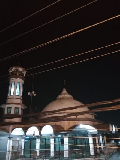A mosque is shown at night in Pandean, Indonesia. The mosque features a dome topped with a crescent moon and a tall, slender minaret with illuminated panels. Overhead, several power lines stretch across the dark sky. The mosque's lower level has an arched facade with visible seating areas behind glass doors and windows.