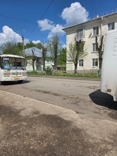 The image shows a street scene in Novomoskovsk, Russia, featuring a white bus with the number "12" displayed. The bus has the license plate "AX 004 71". The street is asphalt with some visible imperfections and patches. A sidewalk is visible on the right side of the image. Buildings with windows are present in the background, along with trees that appear to be freshly in bloom with light green leaves. The sky is bright blue with white, puffy clouds. Power lines are visible stretching across the sky.
