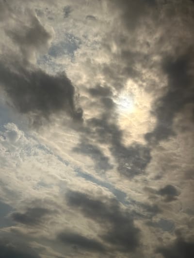 The image shows a sky filled with dramatic, textured clouds, with the sun partially obscured by the dense formations, creating a glowing, ethereal effect. Darker, heavier clouds are interspersed with lighter, wispy ones, and patches of blue sky are visible in the gaps. The lighting suggests either sunrise or sunset, casting a warm, diffused light across the scene. There are no visible people, buildings, or any other identifiable features of a city. The location, Achkhoy-Martan, Russia, is not evident from the visual information presented in the photograph.