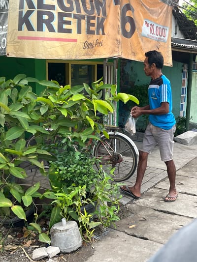A man in a blue t-shirt and grey shorts walks on a sidewalk past a bicycle and a large green bush. The man holds a plastic bag in his right hand. A banner with the words "KRETEK 6" is visible above the man. The ground is paved with concrete slabs. There are buildings in the background.