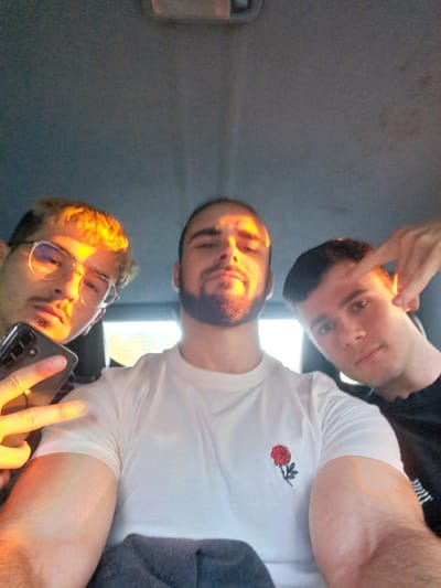 Three young adult males are positioned inside a vehicle in Rahovtsi, Bulgaria, posing for a photograph. The overhead interior of the vehicle, characterized by a dark grey fabric headliner, is visible at the top of the frame. Strong, warm, golden light illuminates the subjects from the left side of the image.

The male on the left, partially framed, wears glasses and has light-colored hair, which appears golden due to the lighting. He holds a dark-colored smartphone in his left hand, with his index and middle fingers extended in a "peace" sign. A metallic ring is visible on his left ring finger. He has a mustache and short facial hair.

The central male, positioned prominently, has dark hair and a beard. He wears a white short-sleeved t-shirt featuring a red rose embroidery on the left chest area. His face is directly illuminated by the golden light, creating distinct highlights and shadows. His right arm, displaying muscular definition, is visible in the lower right portion of the image.

The male on the right, also partially framed, has dark hair and wears a dark-colored t-shirt. His right hand is raised, with his index finger extended upward, pointed towards his temple. His face is partially illuminated by the warm light from the left, casting a portion of his face into shadow. All three individuals appear to be looking towards the camera.