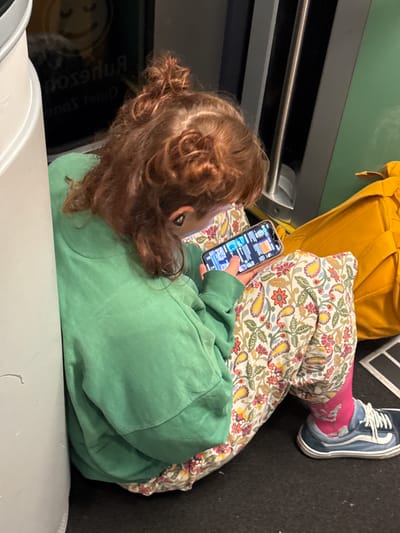 In a public transport setting, possibly a train, a young person with reddish-brown curly hair tied up in a bun is seated on the floor, leaning against a cylindrical object and a green upholstered seat. The person is wearing a green hooded sweatshirt and patterned pants with a floral design, pink socks, and blue and white sneakers. They are engrossed in playing a game on a smartphone, holding it with both hands. A bright yellow bag rests behind them. In the background, a black panel displays text and a smiley face logo. The environment suggests a quiet zone within the public transport. The scene is lit by artificial light, creating a slightly dim atmosphere.