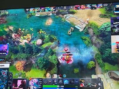 The image shows a screenshot of a video game, likely Dota 2, displayed on a computer monitor. The game features a top-down perspective of a fantasy landscape with a body of water, trees, and terrain. Several in-game elements are visible, including player avatars, health bars, a minimap in the bottom left corner, and various interface elements displaying statistics, item icons, and text. One player is identified by the name "TY, Maladych, YandexMarket" and is engaged in combat with another player. The game's interface indicates a battle is in progress, with scores and timers visible. There are no discernible real-world locations or people present within the image itself, only the digital representation within the video game. Therefore, the location in the city of Sokol'ya Griva, Russia, is not depicted or suggested by the visual content.