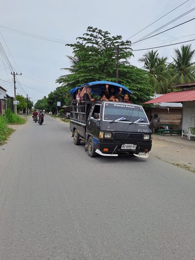 In the city of Cotlagasawa, Indonesia, a black FUSO pickup truck is traveling down a paved road. The truck is carrying several people, both adults and children, on its open bed, which is covered with a blue tarp. Some of the passengers are waving at the camera. The truck has a license plate that reads "BL 8395 KS". In the background, there are trees, palm trees, houses with red roofs, and power lines. Two motorcycles are also visible on the road ahead of the truck. The sky is overcast.