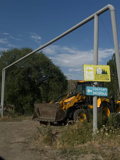 A yellow JCB backhoe loader sits beside a dirt road under a partly cloudy sky. Two signs are fixed to a metal frame. The first sign advertises "KAREN'S cottage Bjni" with information in Armenian, English, and Russian, including a phone number. The second sign, with a blue arrow, points to "ԼՎԱ ԻՆՔԴ МОЙКА," which translates to "Wash It Yourself Car Wash." A 15 km/u speed limit sign is also present. Green trees line the area.