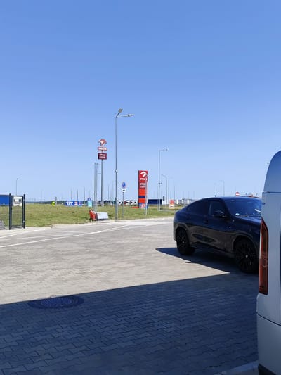 The image depicts an outdoor scene under a clear blue sky, likely at a service station. In the foreground, a dark-colored, modern SUV with black rims is parked on a paved area. To the right, a portion of a silver vehicle is visible. The ground is paved with interlocking grey bricks, with white lines indicating parking or driving lanes. A manhole cover is set into the paving in the lower left quadrant, casting a shadow. In the middle ground, there are several tall lamp posts, some with multiple lights. A prominent red and white sign for "ORLEN stop cafe" is visible, along with other similar signage further in the background. A red trolley with a cleaning attachment is positioned near the grass area. The background features a grassy field and various structures, possibly part of the service station, and a fence on the far left. The general ambiance is one of a bright, sunny day at a roadside establishment.