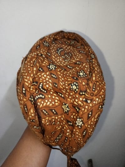 The image displays a brown fabric item with an intricate pattern, held by a human hand, against a plain, light-colored wall in the city of Na Sang, Laos. The fabric features a dense, batik-style design composed of lighter brown, white, and black organic and floral motifs on a dark brown base. The item appears to be folded and gathered, exhibiting multiple creases, with a tie extending from its lower right side. The lower-left quadrant of the image shows a forearm and a portion of a hand, with a medium skin tone, supporting the fabric. A faint shadow is visible on the wall to the right of the fabric item.