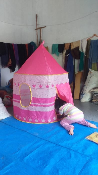 This image depicts an indoor scene located in Cot Madi, Indonesia. The floor is covered by a blue tarp. Against light-colored walls, various garments and textiles are hung on lines and a makeshift wooden support structure. In the foreground, a pink and white castle-themed play tent stands centrally. This tent features a conical roof, alternating bands of pink and white fabric patterned with crowns and dots, an oval window with yellow trim, and an open doorway on its right side. A child, observed from behind, is positioned on hands and knees, barefoot, wearing a multi-striped shirt and light pink pants, with their head and upper torso partially extended into the tent's open doorway. To the right background, a large white sack with blue lettering rests on the floor next to stacks of folded fabric items.