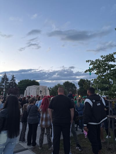 The image, originally rotated 90 degrees clockwise, depicts an outdoor gathering of people, mostly seen from behind, looking towards a distant focal point. The sky, a mix of clear blue and dramatic grey clouds, suggests early evening or late afternoon.

In the foreground, a person wears a black jacket with "CREW" visible in white on the back, holding a pink water bottle. Another individual in a black t-shirt and track pants stands nearby. Further back, a woman with bright red hair is visible, dressed in a plaid shirt and leopard-print trousers, among other casually dressed attendees.

In the mid-distance, a light-colored building or monument stands out, featuring relief carvings that include what appears to be a cross. Green flags or banners hang among the numerous green trees scattered throughout the scene. A wooden fence is partially visible in the background.

The presence of a crowd and "CREW" personnel strongly suggests an organized public event, possibly a ceremony or performance, taking place in Skravena, Bulgaria. The overall mood is one of attentive gathering.