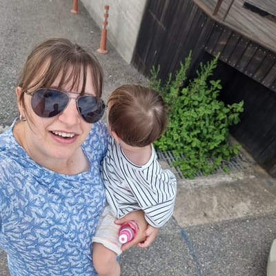 A woman is taking a selfie outdoors while holding a baby. The woman is wearing sunglasses and a blue floral-patterned top. She is smiling, and her hair is blowing in the wind. The baby is wearing a striped shirt and is holding a pink and white toy. The background shows a concrete wall, a wooden structure, and some greenery. The scene appears to be in an urban or semi-urban environment.