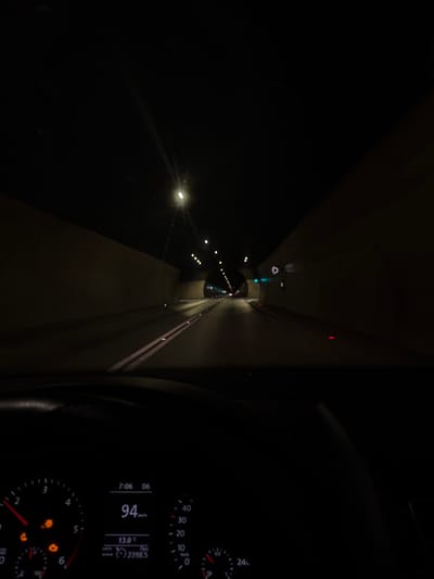 A dark, low-light view from inside a car traveling through a tunnel. The speedometer indicates a speed of 94 km/h. The dashboard clock shows 7:06. A temperature of 13.0 degrees Celsius is also displayed. White tunnel lights recede into the distance. The vehicle is illuminated by these tunnel lights and a reflection of the dashboard lights.