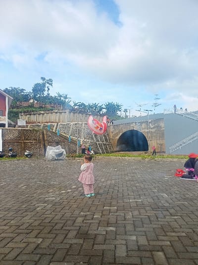 This outdoor photograph captures a young child, appearing to be a toddler, standing centrally on a paved area made of grey interlocking bricks. The child wears a light pink long-sleeved top with a ruffled hem and matching pants, along with light blue footwear, and is looking slightly to the left.
In the mid-ground, a large, bright pink inflatable swan or flamingo-shaped balloon floats in the air, adding a playful element to the scene. The background reveals a multi-tiered landscape with various structures. On the left, part of a building with a red roof and white accents is visible. Below it, a series of stone and concrete retaining walls support a hillside covered in sparse vegetation and trees. To the right of these walls, a substantial concrete structure features a large, dark arched tunnel or underpass opening. Further right, a light grey concrete structure with white stairs ascends towards the top of the embankment. Several adults are scattered in the background; some are near the archway, and others are seated on the ground to the far right.
The sky above is mostly covered with light clouds, indicating an overcast but bright day, likely late morning or early afternoon. The overall atmosphere appears casual and relaxed, suggesting a recreational outing in what seems to be a public park or plaza area. The location context for this image is Kecamatan Pangalengan, Indonesia. No discernible text is visible within the image.
