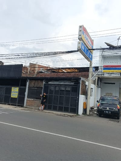 An Indomaret convenience store stands next to a building with a dark metal gate. A man in a black shirt leans against the gate, appearing to drink something. A black van with colorful stripes is parked in front of the store. Power lines run across the top of the frame against a cloudy sky. The foreground is dominated by a wide asphalt road with a white dividing line.