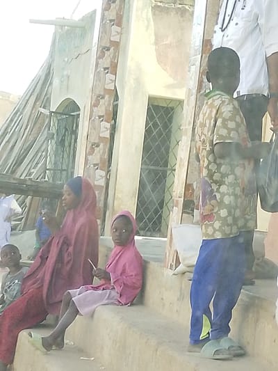 Several children are sitting on the steps of a building. Two girls wear headscarves, one in maroon and the other in pink, with matching attire. A boy stands on the steps to the right, holding a plastic bag. Another boy is seated to the left. A portion of a man wearing a white shirt is visible behind the standing boy. The building behind them has windows and a column with a decorative pattern. Wooden scaffolding leans against the building to the left.
