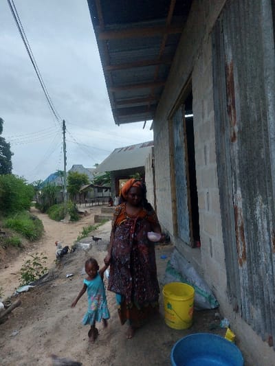 This image depicts a scene on a dirt path in Mwandege, Tanzania, under an overcast sky. In the foreground, an adult female and a young child walk barefoot along the path, holding hands. The adult female, wearing a patterned dress and a headwrap, holds a small white bowl in her left hand. The child, wearing a light blue dress, walks to the left of the adult. To the immediate right of the figures is a building constructed of cinder blocks, with a section of corrugated metal siding and a doorway. A corrugated metal roof overhang extends from the building. Adjacent to the building, a yellow bucket and a blue plastic basin are placed on the ground. Several chickens are visible on the dirt path and near the building. In the midground and background, a wooden utility pole with electrical lines stands to the left of the path, which forks. Further down the path, additional residential structures with corrugated metal roofs and surrounding vegetation are visible.