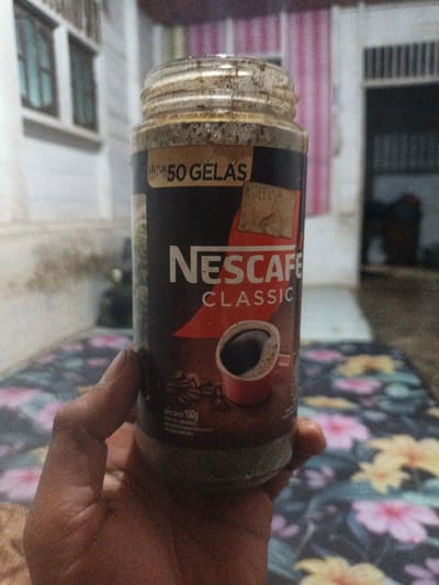 An unidentifiable individual holds a plastic jar of Nescafé Classic coffee in front of a patterned cloth surface. The jar, partially filled with coffee granules, has a label with "UNTUK 50 GELAS" (For 50 Cups) and "NESCAFÉ CLASSIC" printed on it. The background shows a wall with two windows and a doorway, suggesting an indoor setting in a residential area, potentially in Meunasahuket, Indonesia.