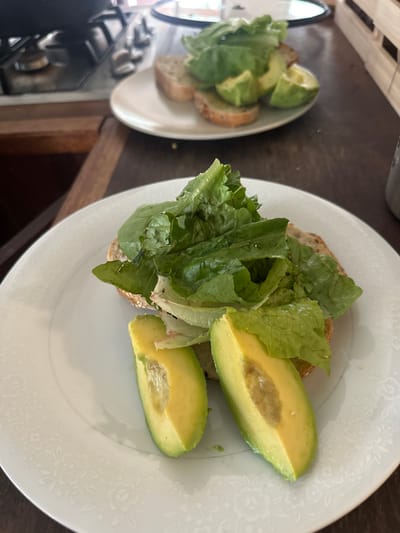 This media file shows a close-up, overhead view of two plates of food on a dark wooden surface. The plate in the foreground contains a sandwich topped with lettuce and slices of avocado. The sandwich appears to be made on whole-wheat bread. The avocado slices are bright green with a light brown pit. The plate has a delicate floral pattern around the rim. To the left, another plate, partially visible, also holds lettuce and avocado on bread. The lighting suggests it is daytime. The overall scene is casual, likely a home setting. No people are visible. There is no visible text.