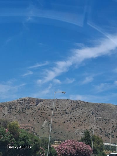 The image depicts a scene in Gazi Municipal Unit, Greece, featuring a steep, rocky mountainside on the right side of the frame. The mountainside is dotted with sparse vegetation and is a predominantly gray-brown color. To the right and slightly below the mountainside, there are patches of green trees and pink flowering bushes. Several streetlights are visible against the backdrop of the mountain. The left side of the frame is dominated by a bright blue sky with wispy white clouds. The photograph appears to have been taken vertically, as the camera branding "Galaxy A55 5G" is displayed along the bottom right edge. The sky is clear, suggesting a sunny day. No people or actions are directly visible in the image.